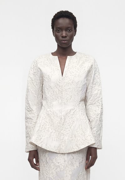 Femme aux cheveux courts et bouclés portant un blazer blanc texturé à manches longues et une jupe assortie, debout devant un fond blanc uni.