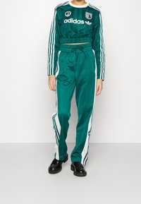 Grön tvådelad träningsdräkt i glänsande tyg, med vita randiga detaljer, en kort topp, elastiskt midjeband och logopatchar. Svarta skor.