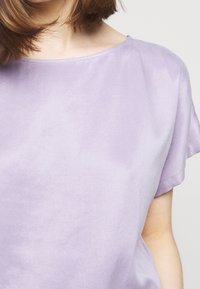 Donna che indossa una maglietta a maniche corte di un leggero colore viola con scollo rotondo, capelli che cadono sulla spalla, su uno sfondo chiaro e semplice.