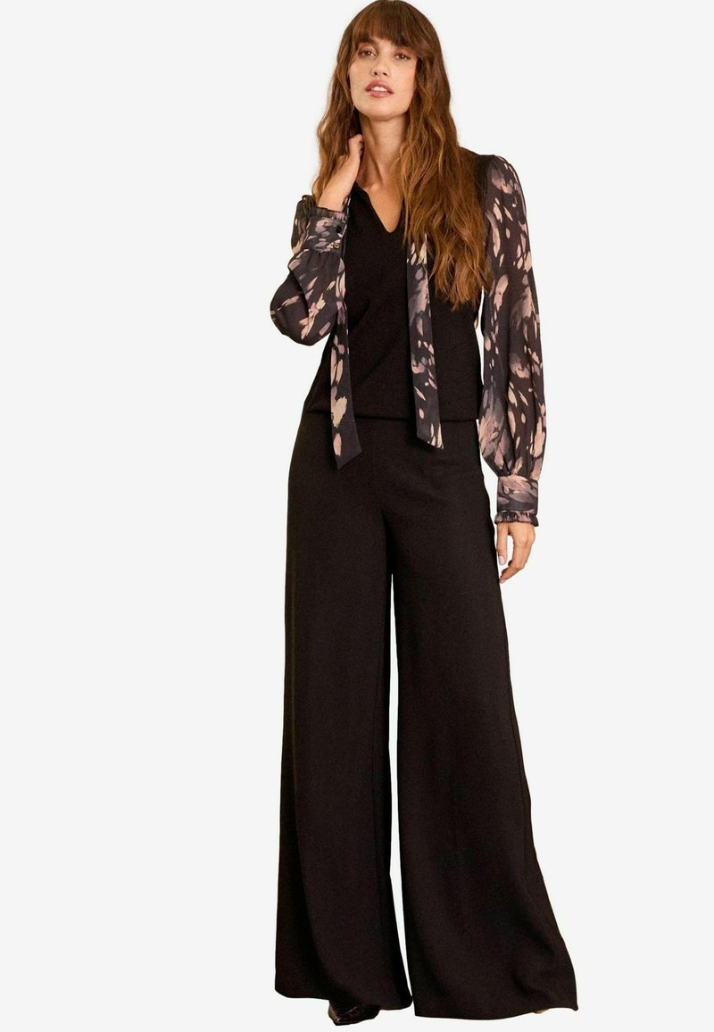 Pantaloni neri a gamba larga abbinati a un top nero a V e a una blusa trasparente a maniche lunghe con motivo, caratterizzata da dettagli floreali in tonalità di rosa e marrone.