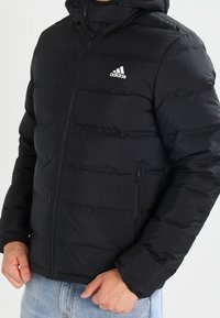 Veste matelassée noire avec capuche, fermeture éclair et poches latérales. Comprend un design matelassé et un petit logo Adidas blanc sur la poitrine.