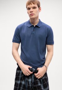 Joven vestido con una camisa polo azul de BOSS, sosteniendo una camisa de cuadros negra y azul atada a la cintura, frente a un fondo blanco liso.