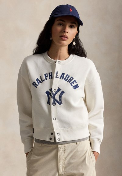 Woman wearing white snap-button sweatshirt with "Ralph Lauren" and blue logo, beige pants, navy cap, and shell earrings, posing with hands in pockets.