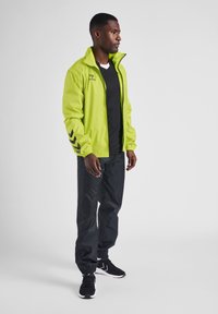 Limegrön jacka med svarta chevronaccentuationer, bärs över en svart skjorta och matchas med svarta byxor och sneakers. Jackan har dragkedjeslutning.