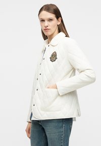 Γυναίκα που φορά λευκό καπιτονέ μπουφάν με έμβλημα στο στήθος και μπλε τζιν, στέκεται με το ένα χέρι στην τσέπη της μπροστά σε λευκό φόντο.