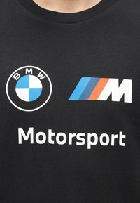 Μαύρο πουκάμισο με λευκό στρογγυλό λογότυπο BMW, μπλε-κόκκινη ριγωτή σήμανση "M" και κείμενο "Motorsport" κάτω από τα λογότυπα στην περιοχή του στήθους.