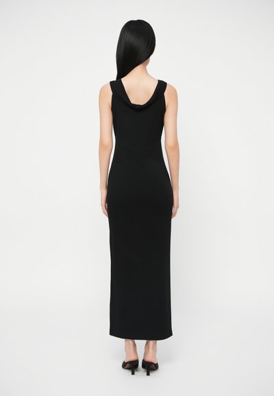 Γυναίκα με μακριά μαύρα μαλλιά, φορώντας μία μαύρη maxi φόρμα χωρίς μανίκια και μαύρα τακούνια, στέκεται με την πλάτη στραμμένη προς την κάμερα.