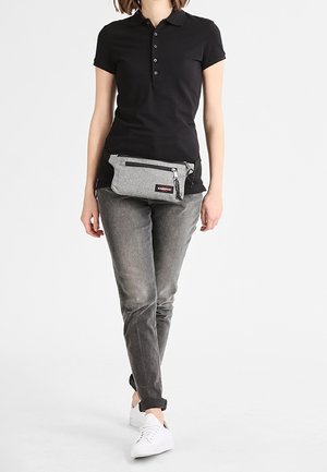 Persona de pie con una camiseta tipo polo negra, jeans grises, zapatillas blancas y una riñonera gris con múltiples cremalleras en la parte delantera.