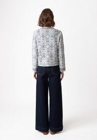 Mėlynas raštuotas švarkas su mandalos dizainu, derinamas su plačiomis tamsiomis džinsinėmis kelnėmis. Apranga pasižymi švaria, modernia forma ir detalėmis.