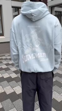 Hoodie azul claro com um bolso frontal, apresentando gráficos brancos de folhas e a palavra "VERÃO" nas costas. Material em mistura de algodão, ajuste descontraído.