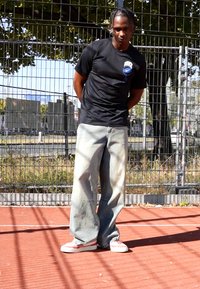 Schwarzes Nike T-Shirt mit Logo, lässige hellblaue Jeans und Sneakers mit roten Akzenten. Steht auf einer roten Asphaltfläche mit einem Zaun im Hintergrund.