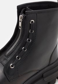 Botín de cuero negro hasta el tobillo con cremallera lateral, herrajes de metal y detalles de cordones. Superficie texturizada con un diseño de suela resistente.