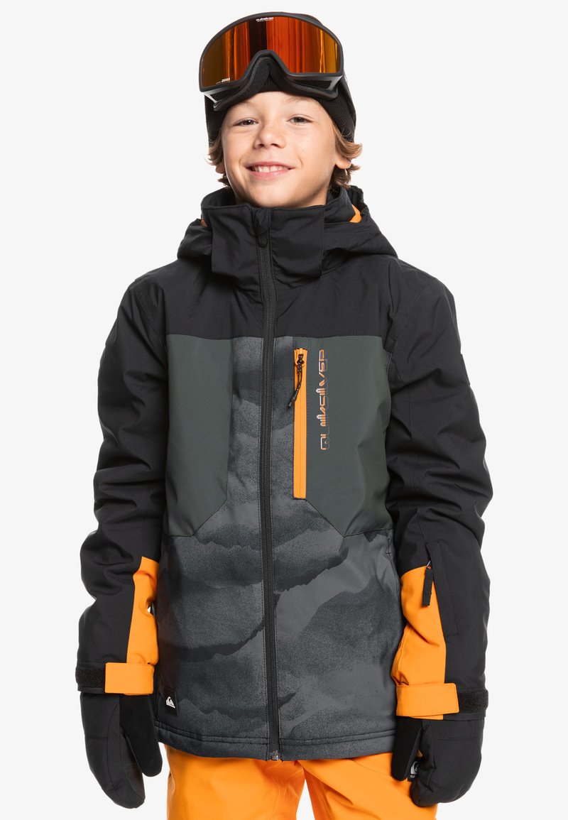 Veste de ski noire et orange avec un motif camouflage gris, poches zippées, poignets réglables et capuche. Portée avec des lunettes de ski.