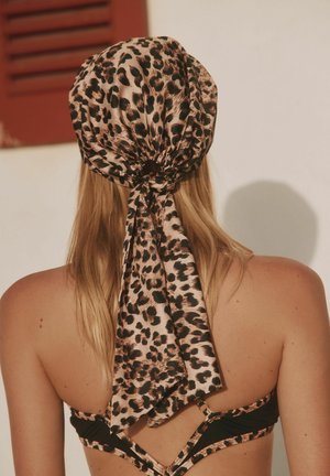 Pikkade blondide juustega naine kannab leopardiprindiga peakatet ja sobivat bikiinikõrgosa, seistes neutraalse taustaga seina ees.