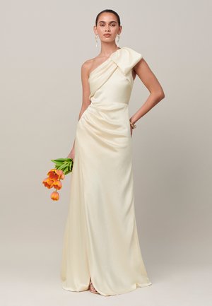 Mujer con un vestido largo de satén crema de un hombro, sosteniendo tulipanes naranjas, de pie frente a un fondo beige liso.