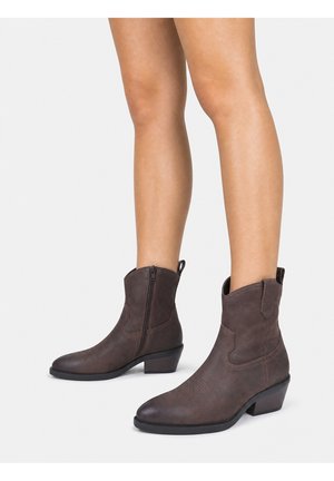 Brune ankel cowboystøvler med sidelås, båret på bare ben mod en ensfarvet lys baggrund.