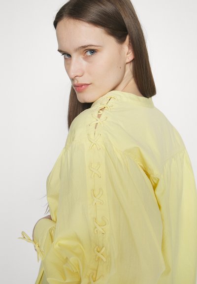 Blouse jaune en tissu léger, avec des détails à lacets aux épaules et des poignets ajustés, offrant une coupe décontractée et une texture douce.