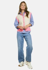 Pletená vesta s růžovými akcenty přes modrobílé pruhované tričko, spárovaná se světle modrými džíny a béžovými sportovními botami.