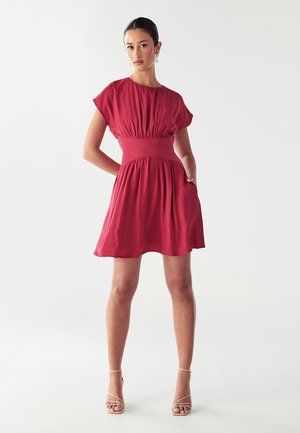 Woman standing with hands in pockets wearing a short-sleeve, knee-length red dress and beige strappy high-heeled sandals against white background.