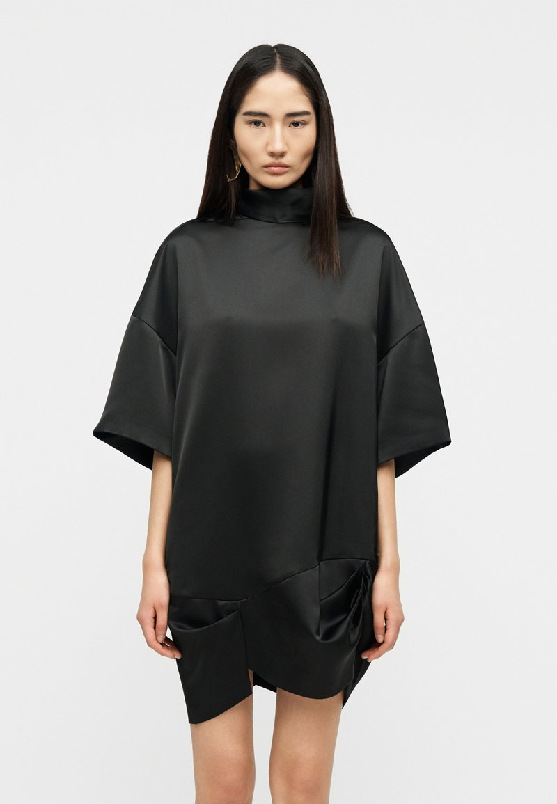 Γυναίκα με ίσια σκοτεινά μαλλιά φοράει χαλαρό μαύρο σατέν φόρεμα με ψηλό γιακά, φαρδιά μανίκια και σούρες στις τσέπες στο εμπρόσθιο στρίφωμα.