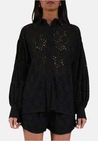Blusa nera all'uncinetto con maniche lunghe a palloncino e chiusura frontale con bottoni. Presenta delicati motivi floreali con una trama aperta e leggera.