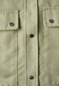 Camisa de pana verde claro con textura de ribetes verticales, dos bolsillos de parche en la parte delantera y botones de presión metálicos.