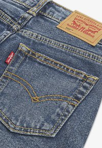 Jean en denim bleu avec une couture jaune, une poche arrière en forme de triangle, et une étiquette en cuir beige avec le logo de la marque.