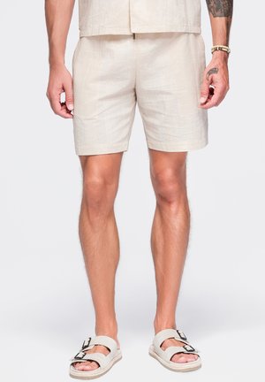 Homme portant un short beige, une chemise à manches courtes assortie et des sandales beige à boucle, debout les bras détendus le long du corps.