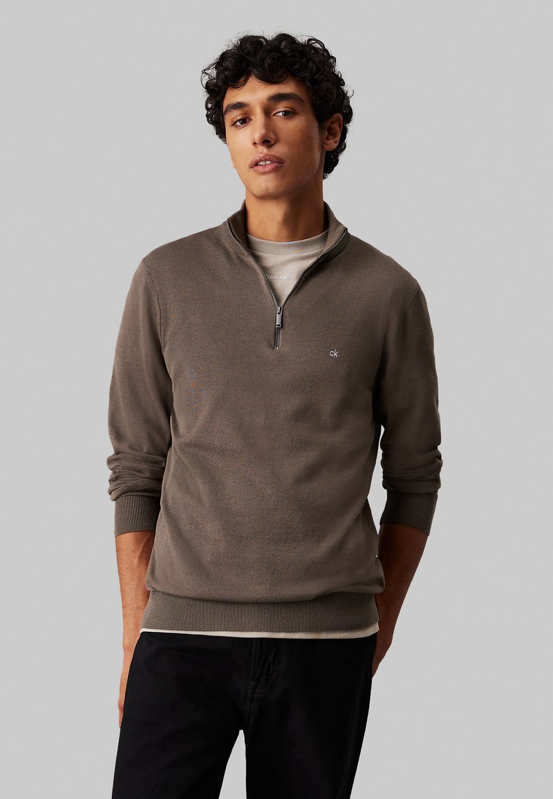 Brauner Reißverschluss-Pullover mit gerippten Bündchen und Saum, der ein kleines Logo auf der Brust zeigt und über einem hellen Hemd getragen wird.