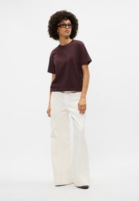 Īsām piedurknēm, bordo krāsas kokvilnas t-krekls, pārī ar augstām, platām baltām biksēm. Modelis stāv pārliecināti pret vienkāršu fona.