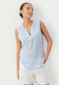 Helles blaues ärmelloses Blusenshirt mit Stehkragen und lässiger Passform, kombiniert mit weißen Shorts. Glattes Material mit leichtem Glanz.