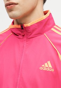Mężczyzna ubrany w różową kurtkę Adidas z pomarańczowymi wykończeniami i logo, zapiętą na zamek, stojący kołnierz, widoczny od podbródka do klatki piersiowej.