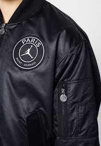 Black bomber jacket featuring a round Paris Saint-Germain patch on the chest, zippered pocket on the sleeve, and ribbed collar detail.