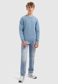 Tiener jongen staat met ontspannen handen, gekleed in een lichtblauwe sweatshirt, vervaagde blauwe jeans en witte hardloopschoenen op een effen achtergrond.