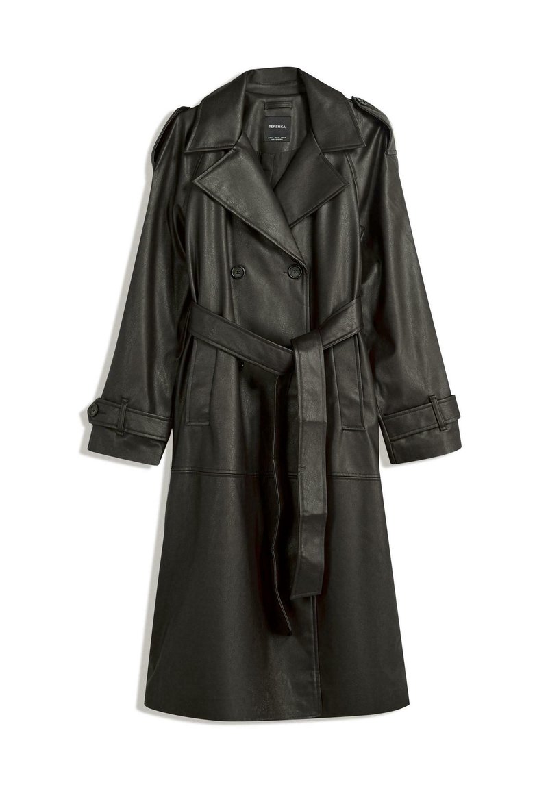 Long trench-coat noir en similicuir avec double boutonnage, larges revers, taille ceinturée et poignets boutonnés sur fond blanc.