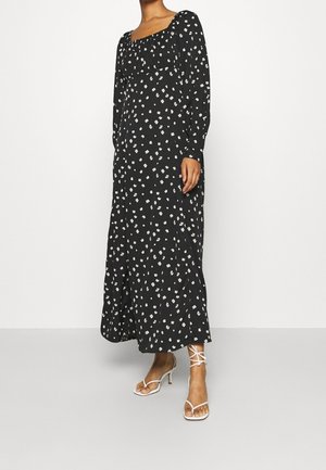 Femme portant une robe maxi noire à manches longues avec un petit imprimé floral blanc et des sandales à talons blanches à brides, debout devant un fond uni.