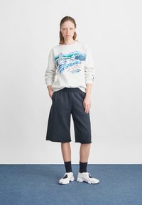 Gai pelēks džemperis ar krāsainu ziemas skatu, kombinēts ar tumši ziliem šortiem un baltiem sporta apaviem. Redzamas vilnas zeķes, teksturēta auduma.