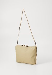 Beige polstret skulderveske med en teksturert overflate, som har et mønstrete stoff, justerbar flette-rem og svarte hardwaredetaljer.