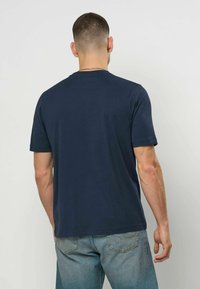 Mann med kort hår iført en ensfarget marineblå t-skjorte med kortermet og lyseblå jeans, stående med ryggen mot en ensfarget bakgrunn.