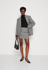 Karierte Blazer- und Rockkombination in Schwarz und Weiß, kombiniert mit einem schwarzen Rollkragenpullover, einer schwarzen Tasche und schwarzen Absätzen. Akzente sind Sonnenbrillen.