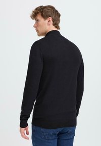 Svart stickad tröja med långa ärmar, hög krage och ribbade mudd och nederkant. Buren över blå denimjeans, sedd bakifrån.