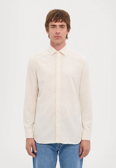 Camisa de manga larga beige con diseño abotonado, cuello clásico y textura suave. Llevada con jeans azules, de pie frente a un fondo blanco.