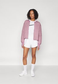 Letvægts lavendel pufferjakke, kort hvid t-shirt med sort logo, hvide shorts, hvide strømper og hvide sneakers.