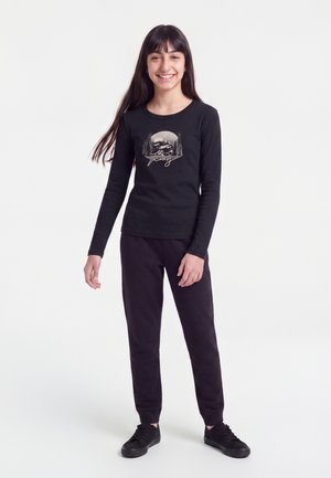Joven niña con cabello largo y oscuro lleva una camisa negra de manga larga con un gráfico de naturaleza y pantalones negros, de pie sobre un fondo blanco.
