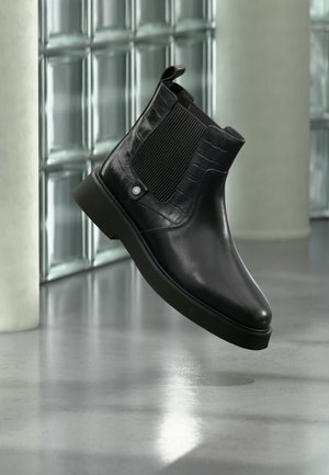 Schwarzer Lederknöchelstiefel mit elastischen seitlichen Einsätzen und strukturiertem Obermaterial, dargestellt vor einem reflektierenden Glasblock und poliertem Boden im Hintergrund.