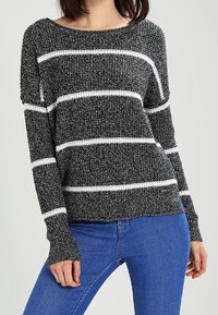 Schwarz-weißer gestreifter Strickpullover mit langen Ärmeln, weitem Ausschnitt und gerippten Bündchen. Getragen über blauen Jeans.