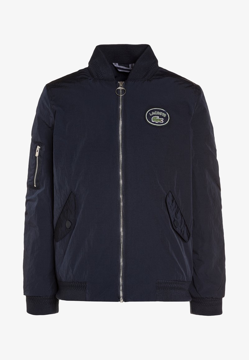 Blouson aviateur bleu marine avec fermeture éclair sur le devant, deux poches à rabat, une poche zippée sur la manche et un patch logo Lacoste sur la poitrine.
