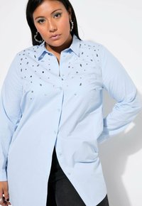 La camicia button-up di colore azzurro chiaro presenta decorazioni metalliche in varie forme sul petto. Maniche lunghe e orlo curvo.