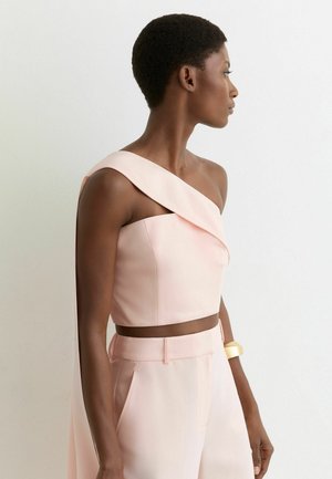 Lühikeste juustega naine, kes kannab heleroosa ühe õlaga lühikest topi ja sobivaid kõrge vöökohaga pükse, seisab profiilis valge seina taustal.