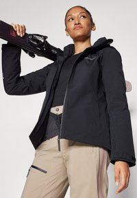 Schwarze Skijacke mit tailliertem Schnitt, Reißverschluss und Kapuze; kombiniert mit beige Hosen, die Taschen und ein pinkes Streifendetail aufweisen.